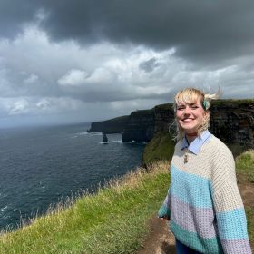 Foto von Dialogidee-Azubi Auli an der irischen Küste. Sie grinst und ihre Haare wehen im Wind.